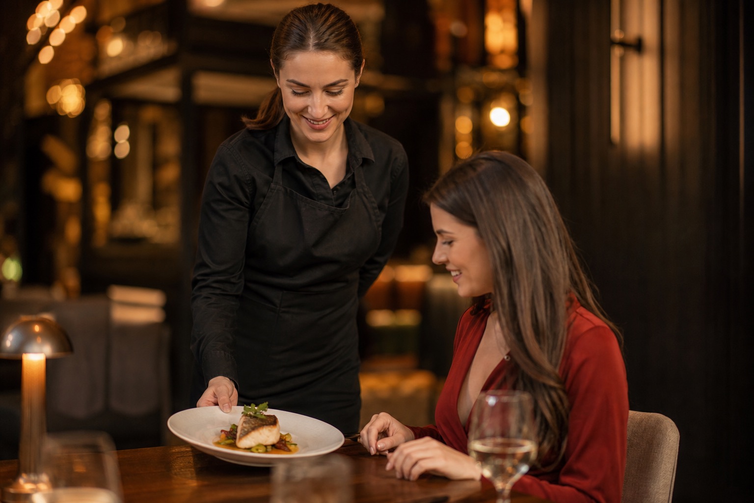 A smiling server bringing a plated dish to a guest in a warmly lit restaurant